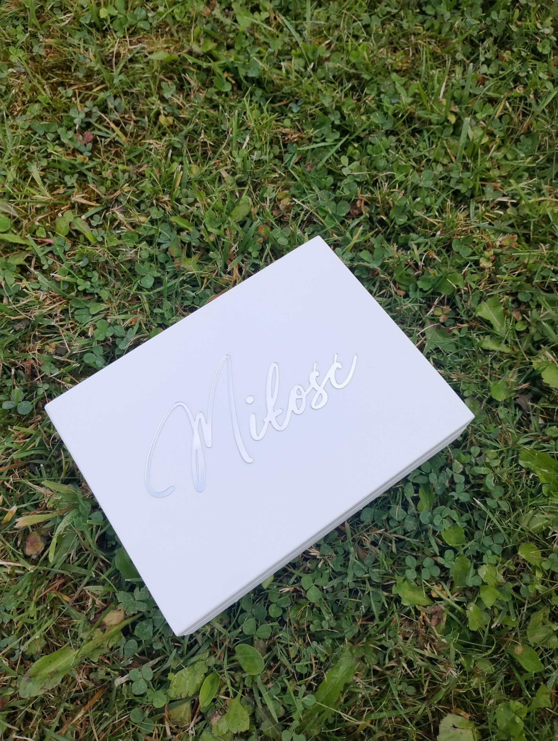 Białe eleganckie pudełko na obrączki z napisem „Miłość” leżące na zielonej trawie