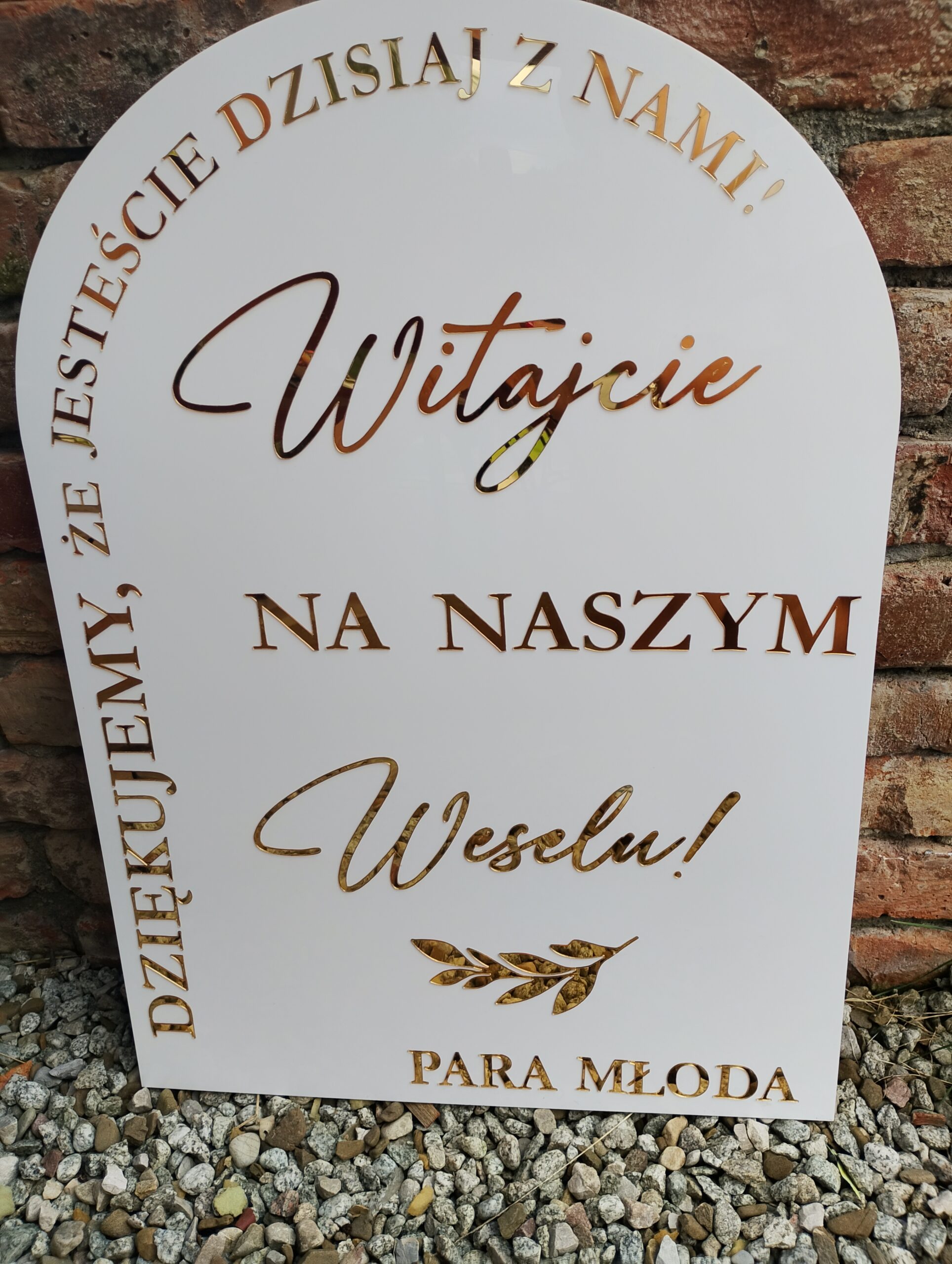 Biała, łukowata tablica powitalna z wypukłymi złotymi literami na tle ceglanej ściany i kamieni.