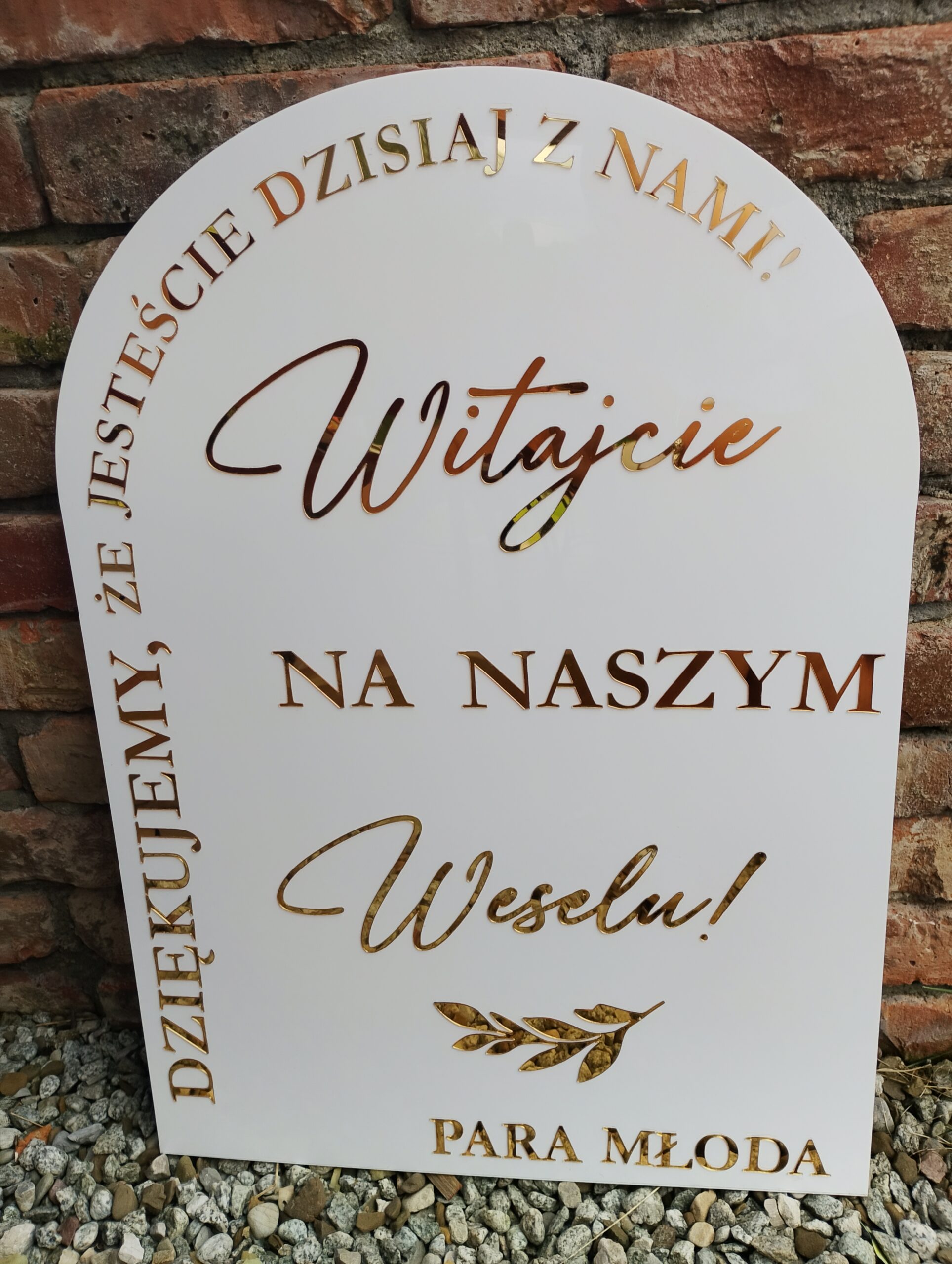 Biała tablica powitalna z napisem "Witajcie na naszym weselu!" i złotymi literami, stojąca na kamykach