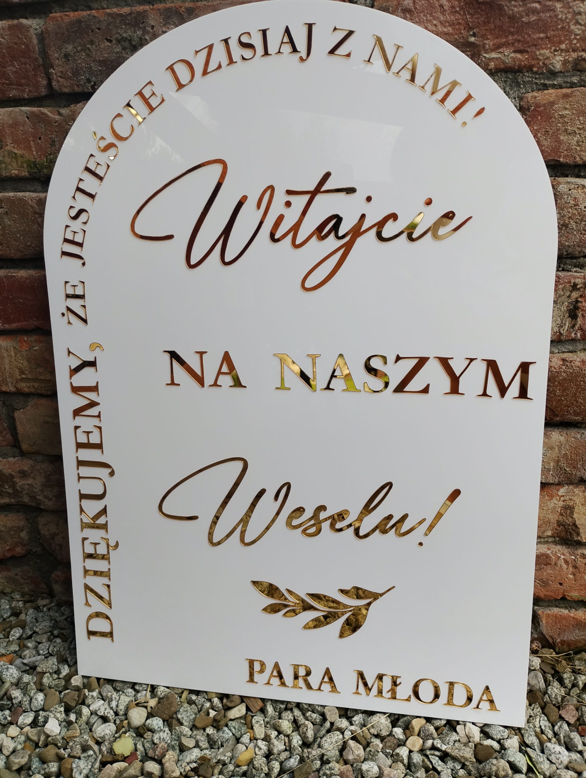 Biała tablica powitalna z napisem "Witajcie na naszym Weselu!" i złotymi literami, stojąca na kamieniach.